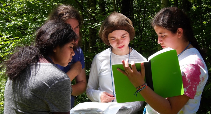 Four students stand in a wooded area and examine printed material. 